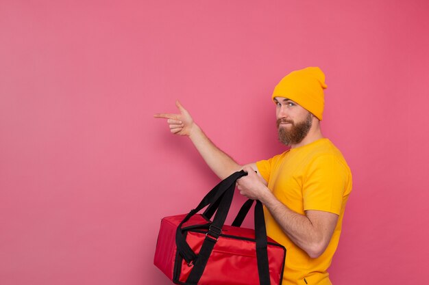 European bearded delivery man with box with food smiling and pointing finger to left on pink