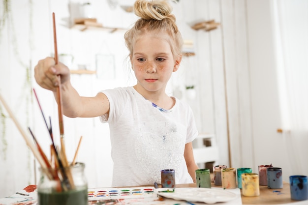 European cute creative child with hair bun snd blue eyes occupied with drawing.