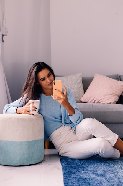 European fit brunette fashion blogger woman sits on floor in living room near sofa