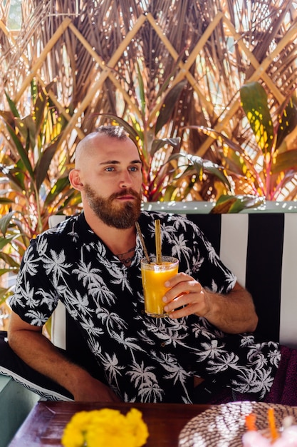 European handsome bearded man drinks mango smoothie in summer cafe, tropical wall behind.