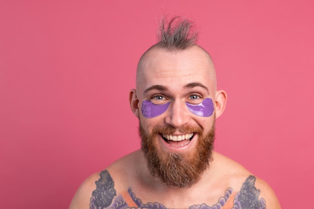 Free photo european handsome bearded tattooed topless man with purple eye patches mask posing to camera on pink