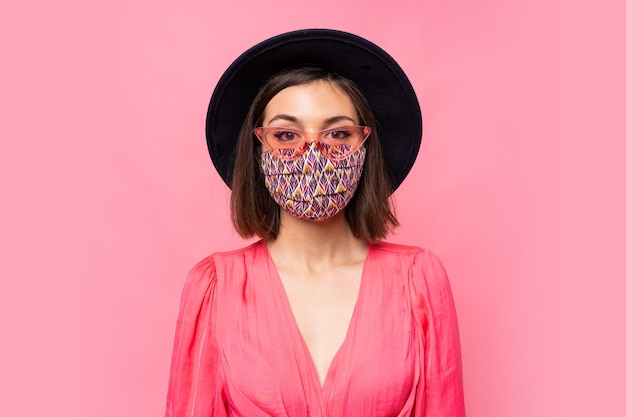 European model dressed protective stylish face mask. Wearing black hat and sunglasses. Posing over pink wall