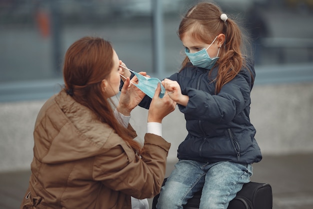 A European mother in a respirator with her daughter are standing near a building.The parent is teaching her child how to wear protective mask to save herself from virus