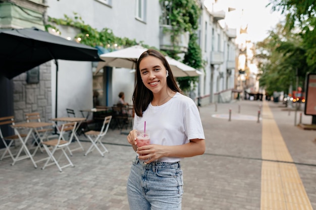 Free Photo european stylish woman with short dark hairstyle wearing white tshirt and jeans posing at camera with wonderful smile and holding smoothie on sunne summer street