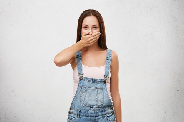 European woman in denim overalls, covering mouth with hand trying to keep silence and not telling secret. Attractive woman keeping secret told by her best friend. Secrecy and conspiracy concept