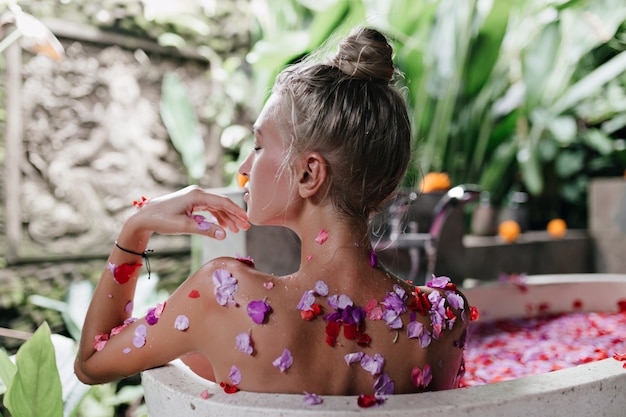 european woman doing spa with flower petals in weekend. Indoor portrait of cheerful woman with tanned skin relaxing in bath with happy face expression.