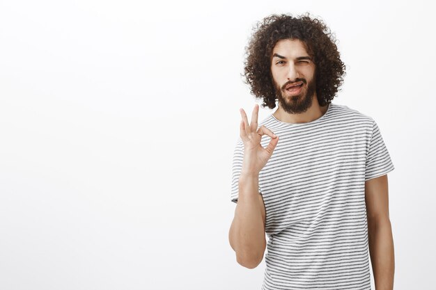 Everything under control babe. Handsome confident hispanic man with beard and curly hairstyle, showing ok or okay sign, winking