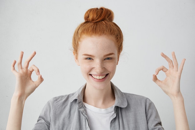 Free photo everything is just fine! cheerful excited young caucasian female with ginger hair knot and freckled skin showing ok gesture with both hands and smiling broadly, enjoying her carefree happy life