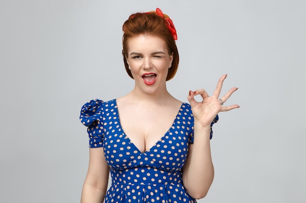 Free photo everything is perfect. isolated picture of elegant gorgeous young ginger female wearing red lipstick and vintage blue dress blinking at camera, keeping mouth wide opened, showing ok gesture