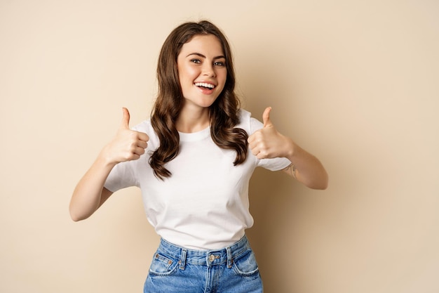 Excellent work. Happy young woman showing thumbs up in approval and smiling, standing in t-shrit over beige background.