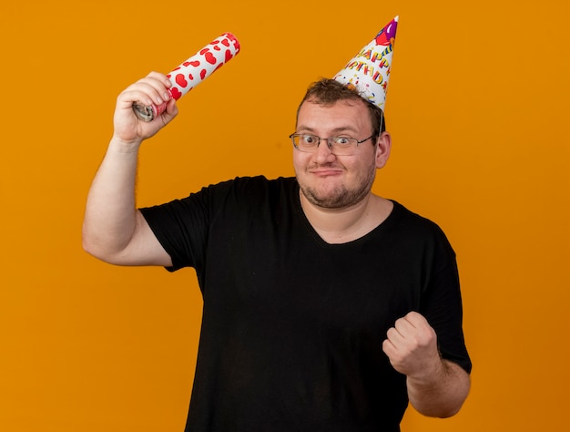 Free photo excited adult slavic man in optical glasses wearing birthday cap holds confetti cannon and keeps fist