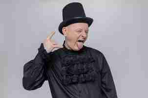 Free photo excited adult slavic man with top hat and in black gothic shirt stucks out tongue and gestures horns sign