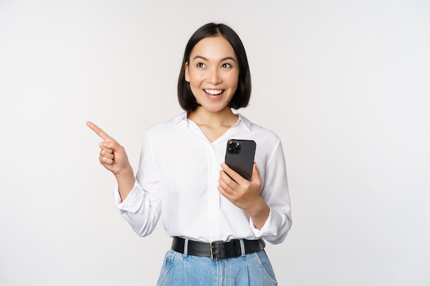 Free photo excited asian girl shopping on mobile app holding phone and pointing finger left at empty copy space showing promo text standing with smartphone against white background