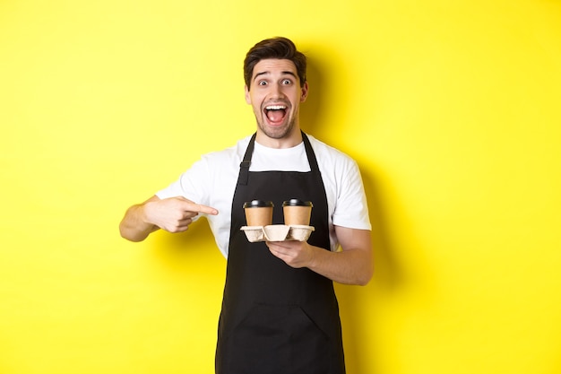 Excited barista in black apron pointing fingers at takeaway coffee cups, standing against yellow background happy.