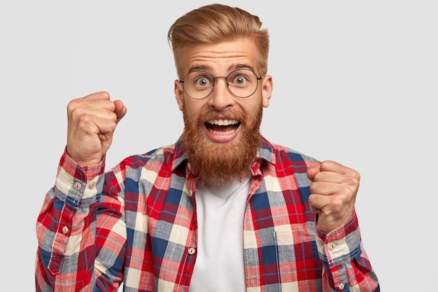 Free Photo excited bearded young ginger man has joyful expression, clenches fists, being in good mood as celebrates his victory