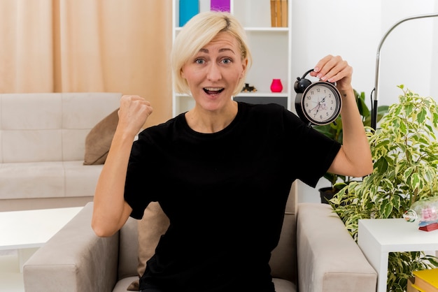 Free Photo excited beautiful blonde russian woman sits on armchair keeping fist and holding alarm clock inside the living room