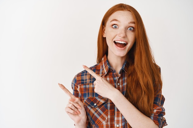 Excited beautiful girl checking out promo offer, pointing at logo and smiling amazed, standing against white wall