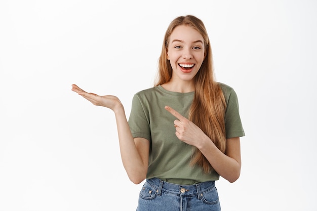 Excited blond girl, smiling, pointing at open hand as if showing at item in palm, recommending product, display advertisement, standing against white wall