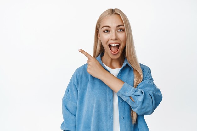 Excited blond woman smiling, pointing finger at upper left corner with amazed face, showing awesome news, big sale banner on white