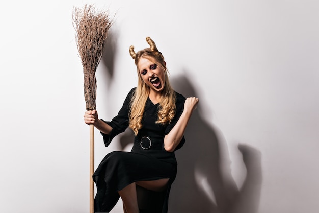 Free Photo excited blonde witch posing with broom. beautiful vampire in black dress expressing happiness in halloween.