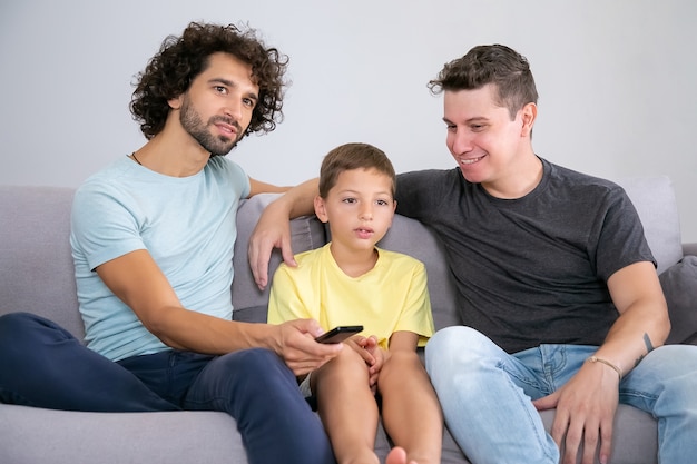 Excited boy and his cheerful dads watching TV show at home, sitting on couch in living room, hugging, using remote control, looking away. Family and home entertainment concept