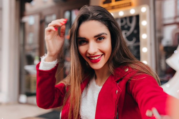 Free photo excited brown-haired girl with stylish makeup making selfie on blur urban space