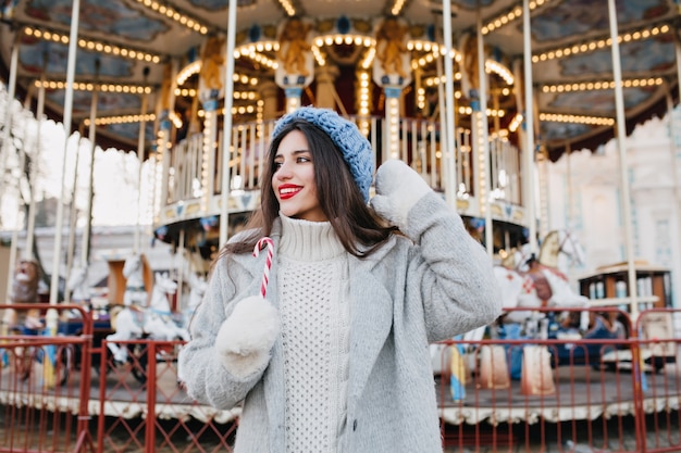 Free photo excited brunette girl in blur knitted hat waiting for friend in amusement park in winter day. outdoor photo of happy woman with dark hair holding candy cane and posing in front of carousel.