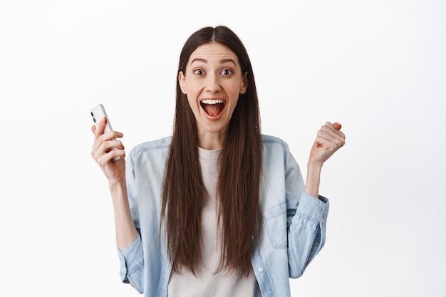 Excited brunette girl screams amazed, holding smartphone and celebrating, winning online, receive great news, achieve app goal, standing over white wall