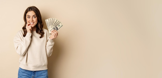 Free Photo excited brunette woman holding money and biting finger with yearning face watns to buy smth shopoholic with cash standing over beige background