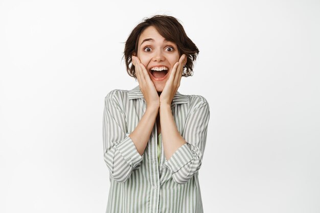 Excited brunette woman laughing and smiling happy, receive surprise, look with happy and amazed face at camera, standing in shirt over white background.