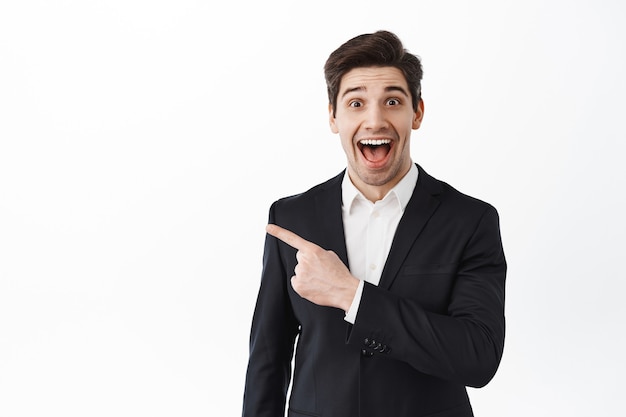 Excited business man, corporate entrepreneur in suit gasping amazed, pointing aside at copyspace with impressed smiling face, checking out awesome deal, white wall