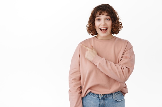 Excited caucasian girl gasp, say wow and pointing at upper left corner with surprised and impressed face, checking out advertisement, amazing news, standing over white background.