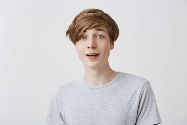 Excited caucasian young man with braces wears gray t-shirt watches football match with friends, being involved in game, looks  happily because of scored goal, rejoices team`s victory