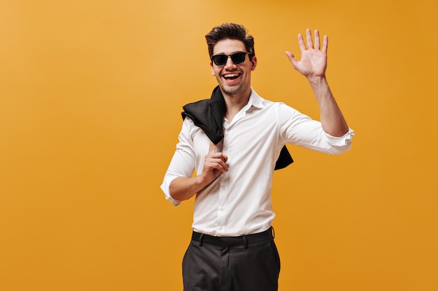 Free photo excited charming brunet man in white shirt, sunglasses and black pants smiles, holds jacket and waves hand in greeting on orange wall.