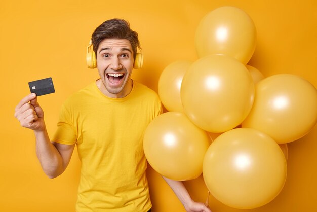 Excited cheerful adult man glad to get money on bank account listens music via wireless headphones holds bunch of inflated balloons wears casual clothes isolated over vivid yellow background