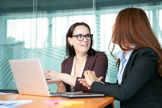 Excited cheerful businesswomen discussing project while sitting at open laptop