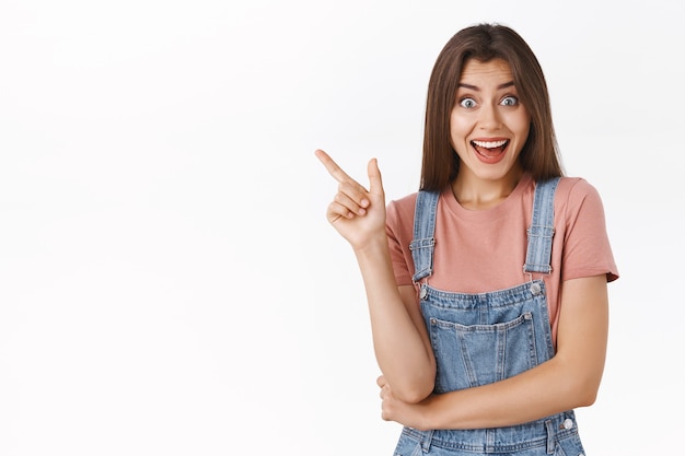 Excited cheerful cute modern girl in overalls, t-shirt pointing upper left corner, smiling amused discuss with girlfriend amazing promotion, giveaway, standing white background thrilled