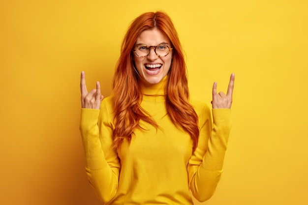 Free photo excited cheerful redhead woman shows rock n roll gesture makes horns with fingers laughs joyfully enjoys listening favorite rock music wears casual turtleneck.