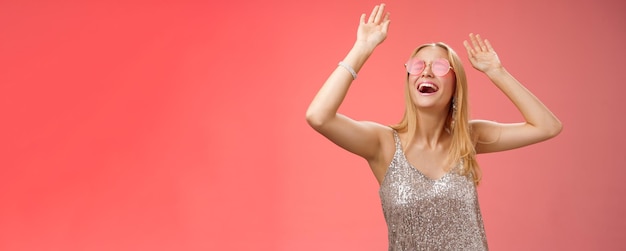 Free photo excited chilling energized young blond woman in silver stylish glittering dress sunglasses raise hands up having fun dancing dancefloor nightclub throw party celebrate bday red background