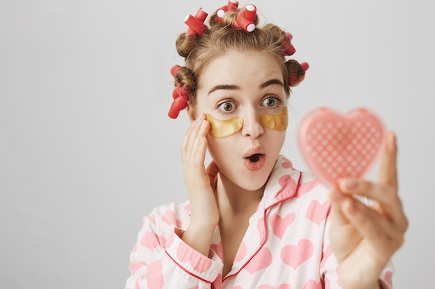 Free photo excited cute girl in hair curlers and eye patches looking in mirror