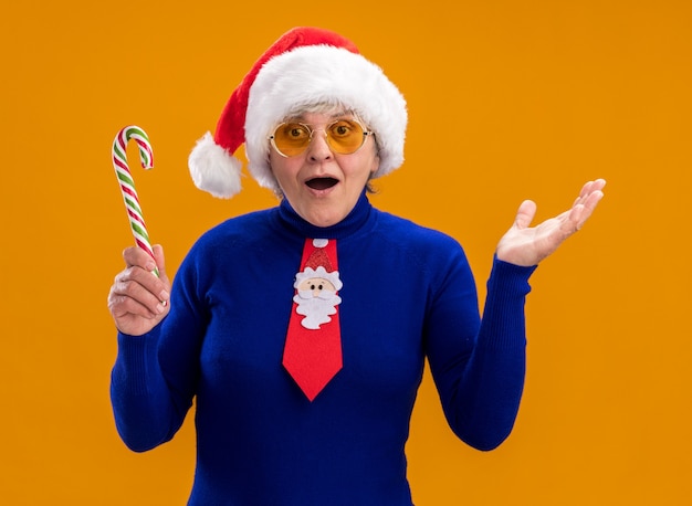 Free Photo excited elderly woman in sun glasses with santa hat and santa tie holds candy cane and keeps hand open isolated on orange wall with copy space