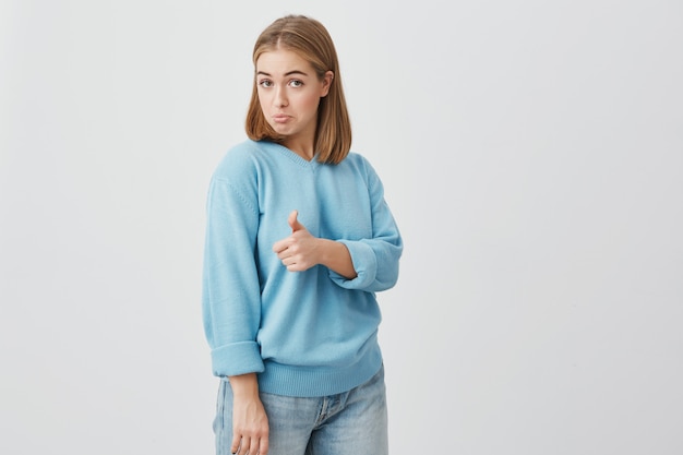 Excited emotional young European female customer with fair hair dressed in blue clothes giving her thumb up, showing how good a product is. Gestures and body language