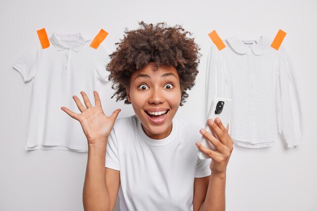 Excited emotional young woman with cheerful expression keeps palm raised reacts happily on amazing news uses mobile phone dressed casually isolated over white background with clothes behind.