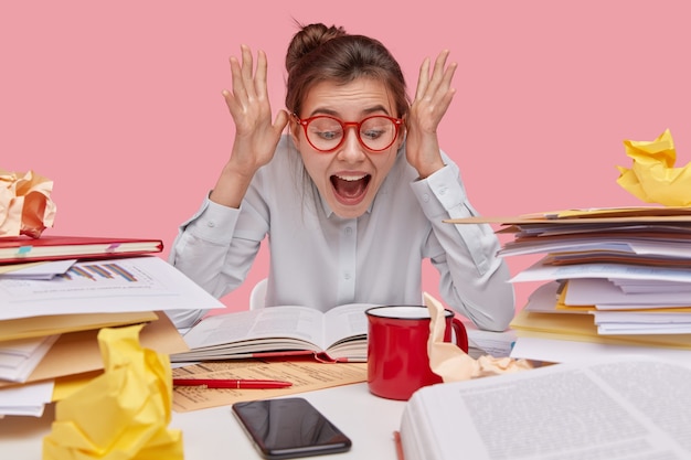 Excited emotive woman raises hands near face, keeps mouth widely opened, reacts on astonishing information written in book, wears red rim glasses