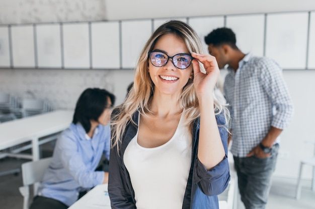 Excited european female student holding glasses and posing between lectures. Indoor portrait of smiling woman standing beside asian and african university mates during seminar.