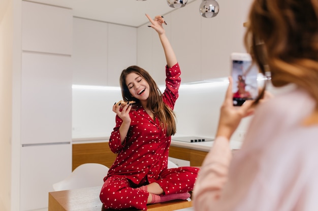 Excited european girl eating pizza during home photoshoot. Adorable woman in red sleepwear sitting on table while her sister taking pictures with phone.