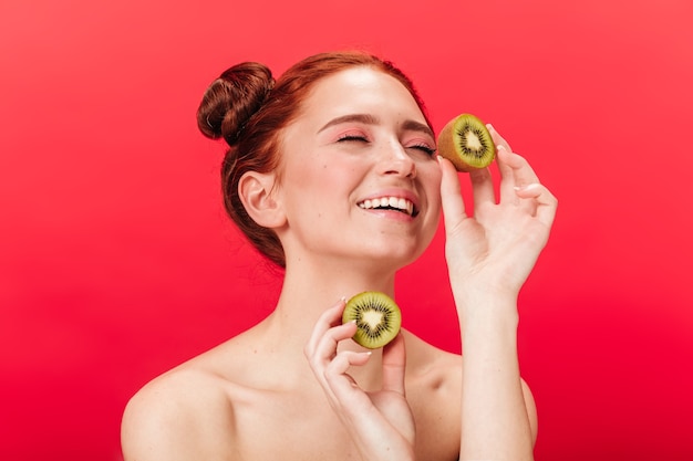 Free photo excited european girl holding kiwi. studio shot of carefree woman with exotic fruits isolated on red background.