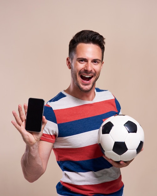 Free photo excited fan holding soccer ball and showing mobile phone