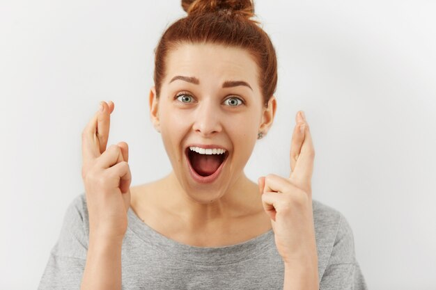 Excited and fascinated young Caucasian female with ginger hair keeping fingers crossed for good luck