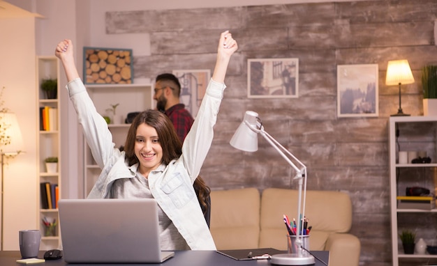 Free photo excited female freelancer with hands raised after reading a good news on laptop. boyfriend talking on the phone in the background.